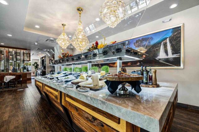 Upscale restaurant buffet display with a marble and wood counter, assorted dishes, crystal chandeliers, and a large landscape artwork on the wall.