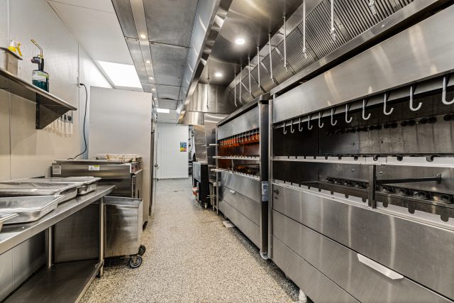 Narrow commercial kitchen corridor lined with stainless steel equipment, ventilation hoods, prep surfaces, and refrigerated drawers.