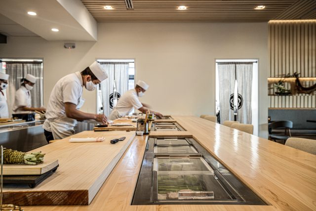 Sushi chefs in white uniforms and masks preparing food behind a light wood omakase counter with built-in refrigerated display cases.