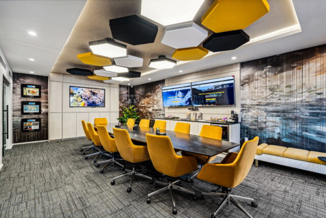 Modern corporate conference room with yellow upholstered chairs, hexagonal acoustic ceiling panels, dual wall-mounted displays, and custom wall graphics