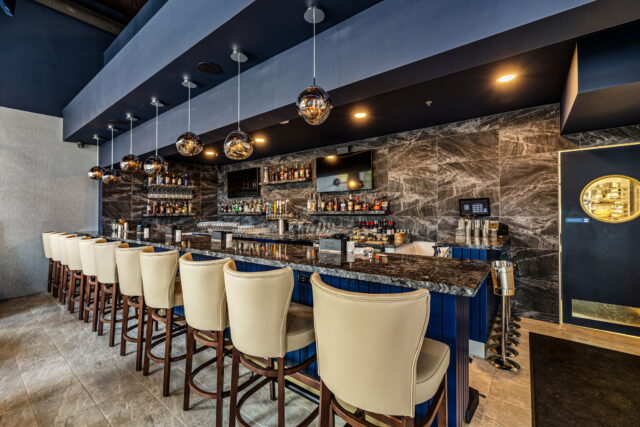 Upscale bar interior with cream upholstered bar stools, navy blue bar front, dark marble surround, full back bar, and globe pendant lighting
