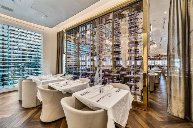 Upscale fine dining restaurant interior with white linen tables, floor-to-ceiling glass wine wall, and herringbone hardwood floors.