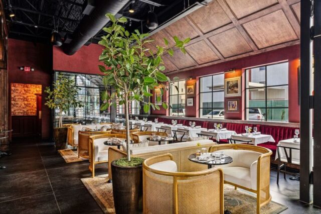 Fine dining restaurant interior featuring cane lounge chairs, white-tablecloth seating, rich red walls, and an indoor fig tree