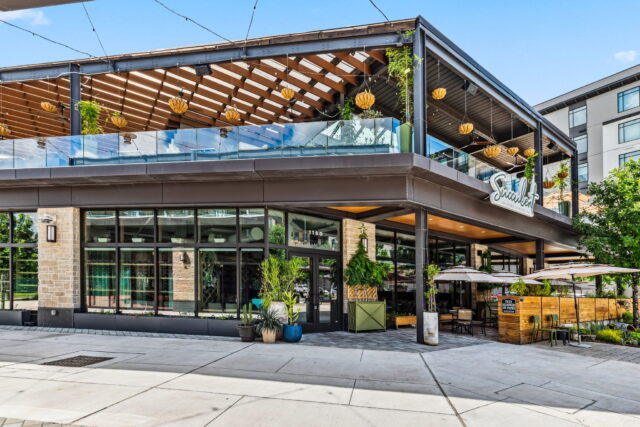 Exterior of a modern two-story restaurant with a rooftop pergola, hanging lanterns, lush greenery, and an outdoor patio with umbrellas