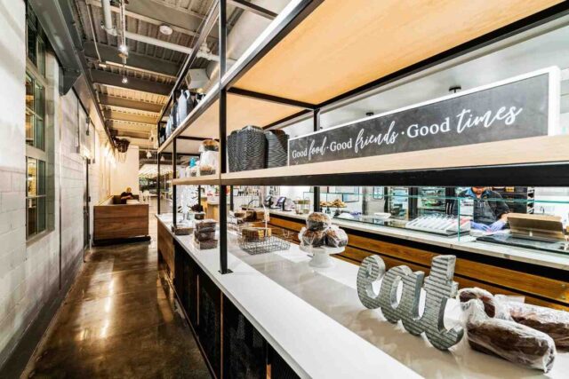 Cafe and bakery interior with open steel shelving, baked goods display counter, polished concrete floors, and an open kitchen